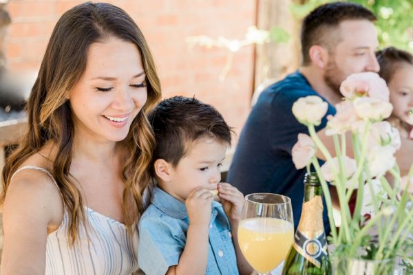 A family enjoys a meal outdoors. The woman smiles, holding a child. A man and another child are in the background with flowers on the table.