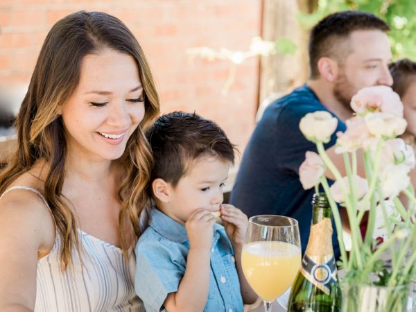 A family enjoys a meal outdoors. The woman smiles, holding a child. A man and another child are in the background with flowers on the table.