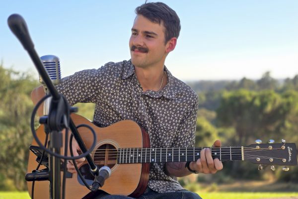 A person sitting outdoors playing an acoustic guitar, with microphones set up, enjoying a scenic view.