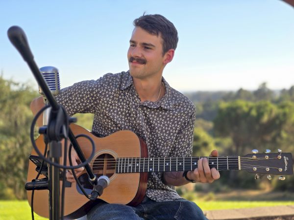 A person sitting outdoors playing an acoustic guitar, with microphones set up, enjoying a scenic view.