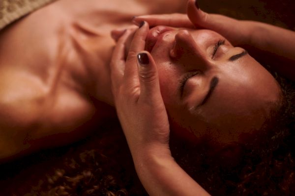 A person receiving a relaxing facial massage with two hands gently placed on their face, eyes closed, and covered by a towel at the chest.