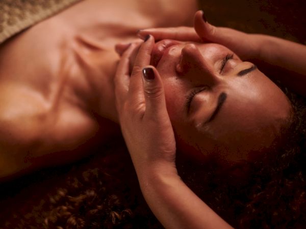 A person receiving a relaxing facial massage with two hands gently placed on their face, eyes closed, and covered by a towel at the chest.