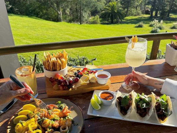 A scenic outdoor table with tacos, seafood, fries, drinks, and a view of a lush green lawn and trees under a clear sky.