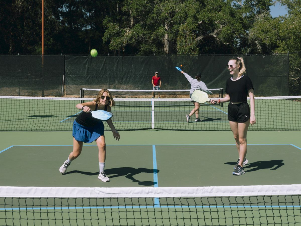 People playing pickleball on a sunny day. One person serves while others are ready to play, with trees in the background.