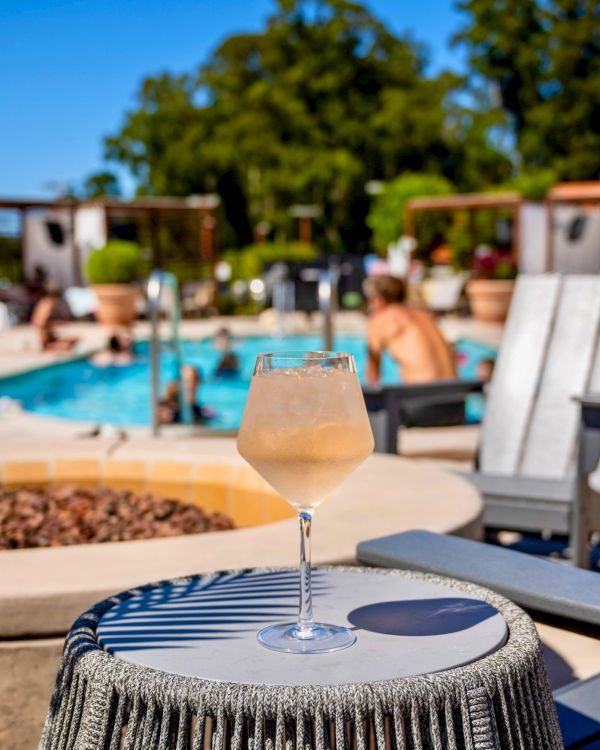 A glass of chilled beverage sits on a table by a pool, with people relaxing in the background on a sunny day.