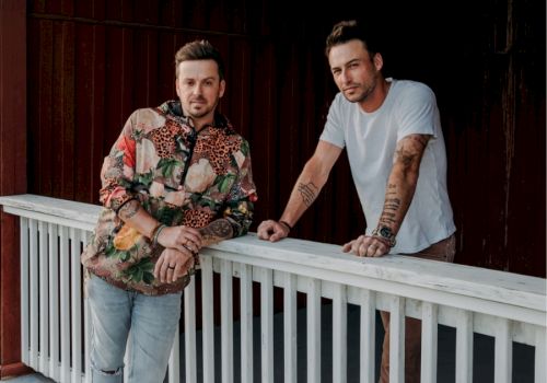 Two people are leaning on a white railing against a dark wooden backdrop.