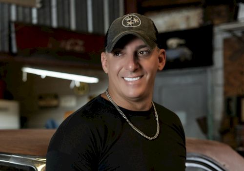 A person wearing a black shirt, chain necklace, and cap is smiling, standing in an indoor setting with visible industrial elements.