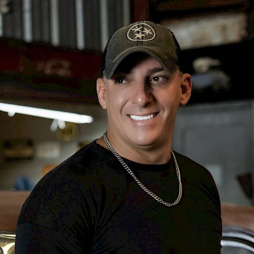 A person wearing a black shirt, chain necklace, and cap is smiling, standing in an indoor setting with visible industrial elements.