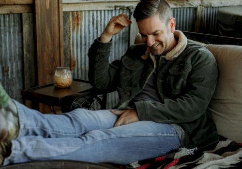 A man is relaxing on a couch by a fire pit, wearing a jacket and jeans, with a drink on a small table beside him.