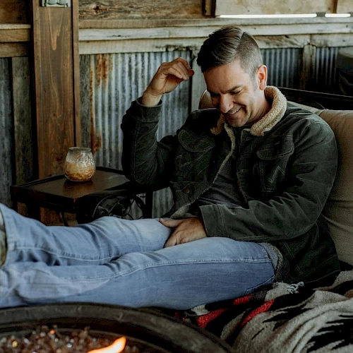 A man is relaxing on a couch by a fire pit, wearing a jacket and jeans, with a drink on a small table beside him.