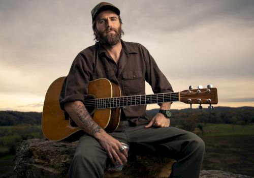 A person with a guitar sits on a rock, wearing a cap and holding a drink, set against a rural landscape backdrop under a cloudy sky.