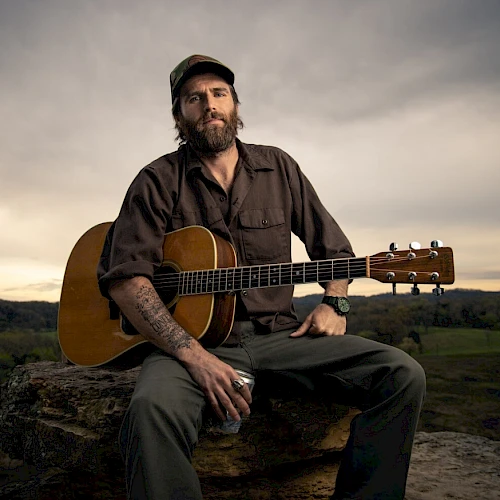 A person with a guitar sits on a rock, wearing a cap and holding a drink, set against a rural landscape backdrop under a cloudy sky.