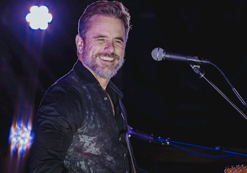 A man is performing on stage with a guitar, smiling at the audience, under bright stage lights, with a microphone nearby.