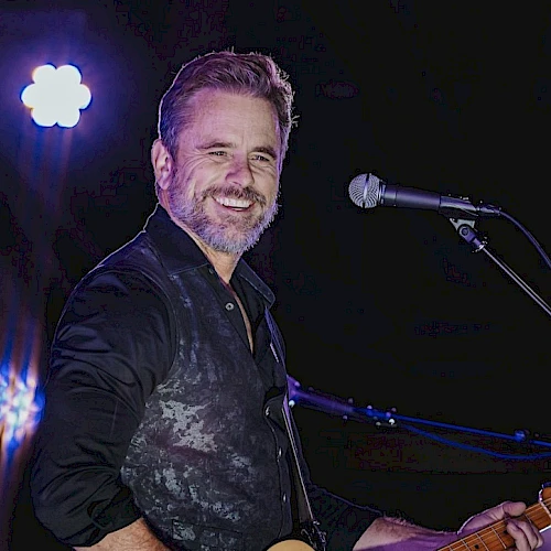 A man is performing on stage with a guitar, smiling at the audience, under bright stage lights, with a microphone nearby.