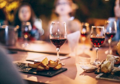 The image shows a table with several glasses of red wine, plates of food, and people in the background, slightly out of focus.