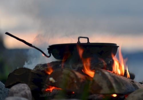 A pot over an open flame, surrounded by rocks, with smoke rising against a blurred outdoor backdrop.