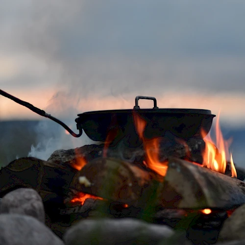 A pot over an open flame, surrounded by rocks, with smoke rising against a blurred outdoor backdrop.