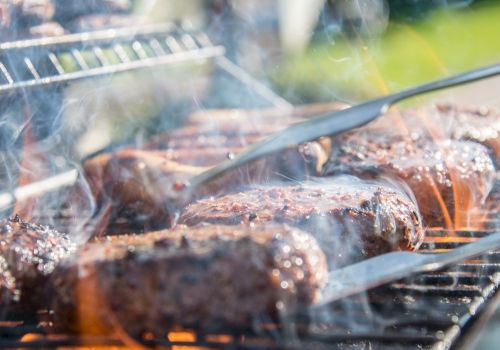Grilled meats are being cooked on a barbecue with metal tongs in use, surrounded by smoke and flames.