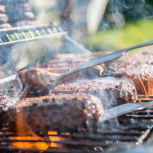 Grilled meats are being cooked on a barbecue with metal tongs in use, surrounded by smoke and flames.