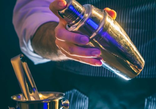 A person holding a shiny cocktail shaker above an ice bucket, possibly preparing a drink.