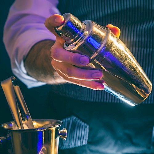 A person holding a shiny cocktail shaker above an ice bucket, possibly preparing a drink.