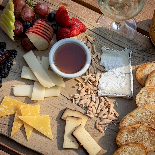 A charcuterie board with assorted cheeses, fruits, nuts, bread slices, and a dip, accompanied by a glass of white wine on the side.