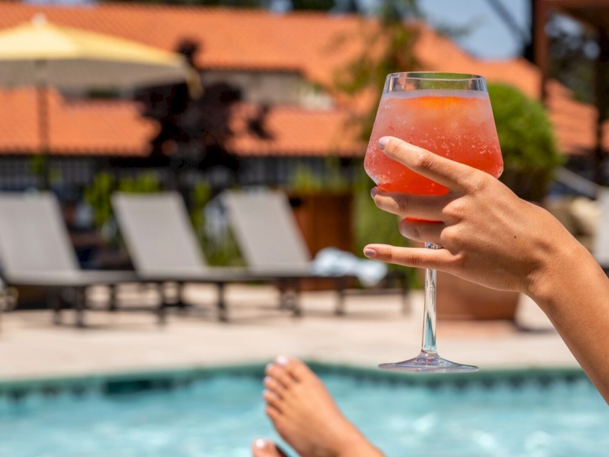 A person relaxes by a pool, holding a chilled glass of red drink, with loungers and an umbrella in the background.