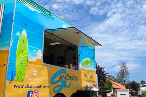 A colorful food truck named "Sandbox," with surfboards painted, set near a pool area. The sky is partly cloudy.