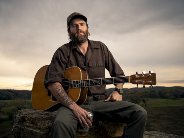 A person with a beard and cap holds an acoustic guitar, sitting outdoors on a rock with a scenic backdrop.