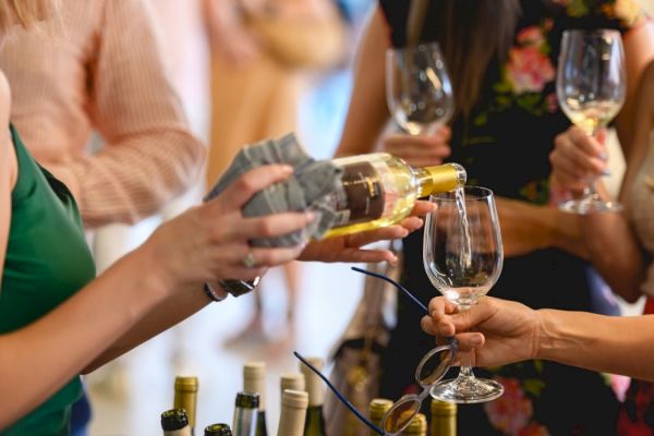 People at a social gathering are pouring white wine into glasses. Bottles are in the foreground.