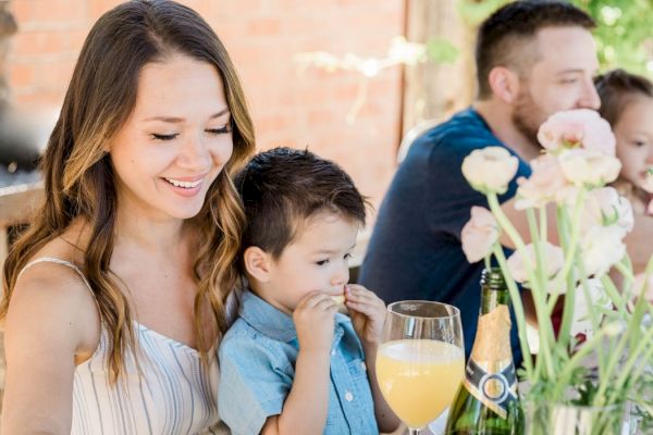 A woman and child are seated at a table with flowers, sharing a joyful moment, with drinks and another adult visible in the background.