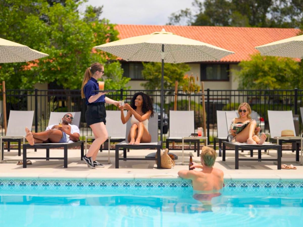 People relax by a pool under umbrellas; some lounge while one stands, possibly a server. A person is in the pool in the foreground.