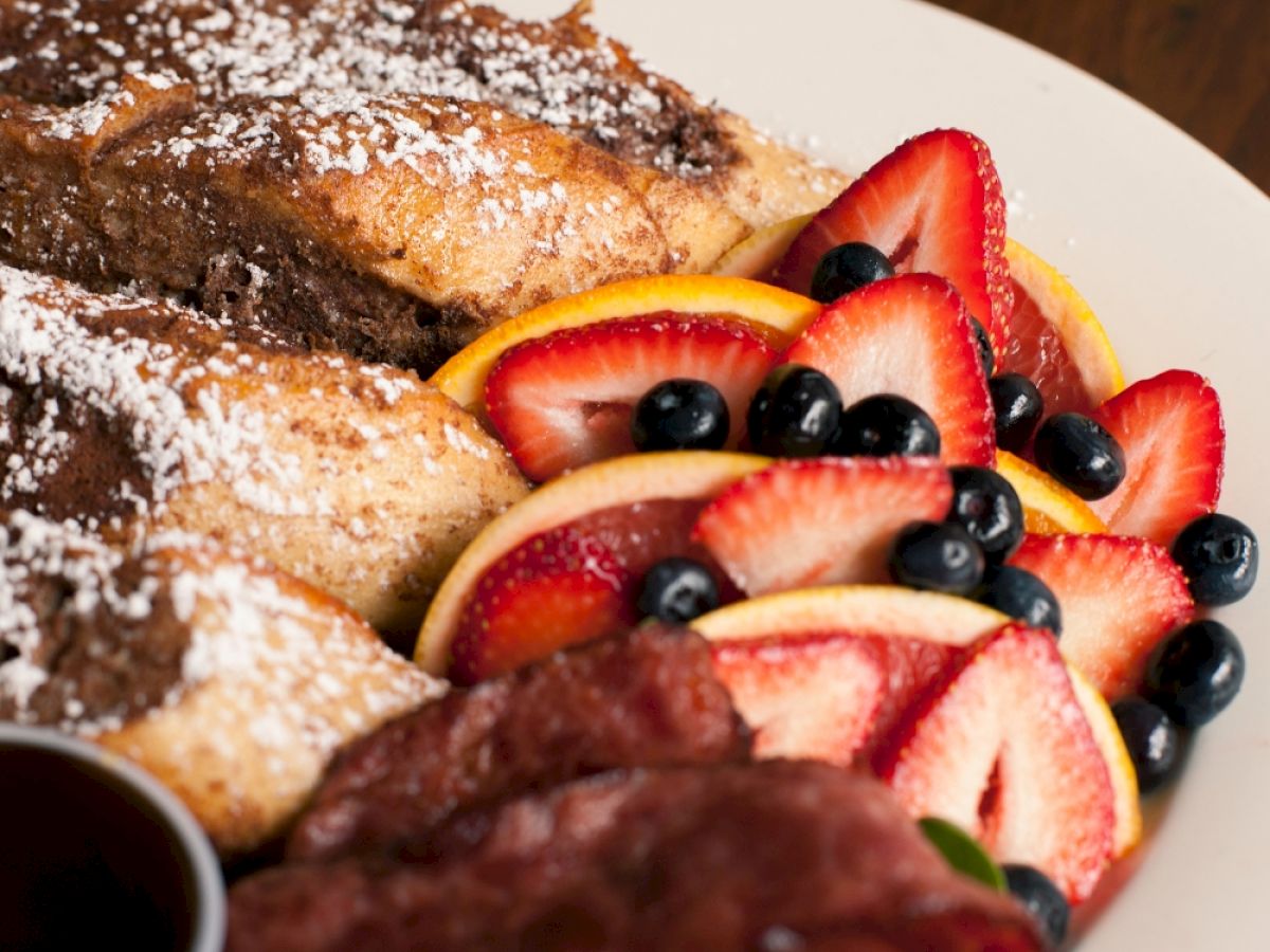 The image shows French toast with powdered sugar, sliced strawberries, blueberries, and citrus slices on a plate.