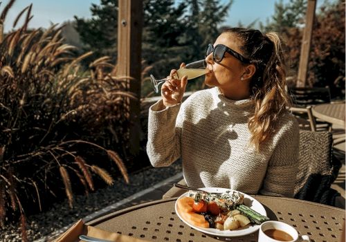 A woman in sunglasses is enjoying a drink outdoors, seated at a table with a plate of food, surrounded by greenery and sunlight.