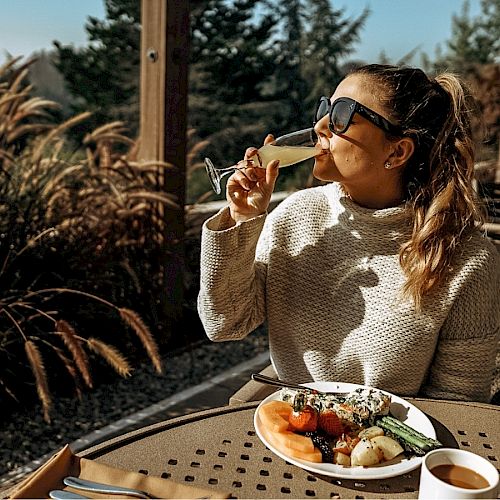 A woman in sunglasses is enjoying a drink outdoors, seated at a table with a plate of food, surrounded by greenery and sunlight.