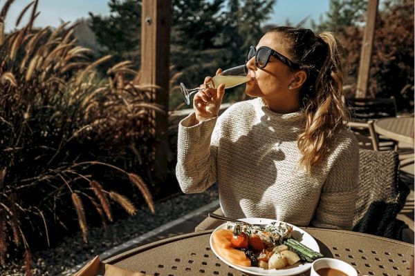 A person in sunglasses is outdoors, enjoying a drink with a plate of food and a cup, surrounded by greenery and sunlight.