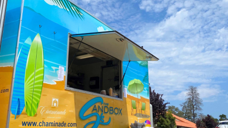 A colorful food truck named "Sandbox," with surfboards painted, set near a pool area. The sky is partly cloudy.