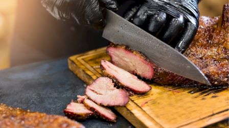 A person wearing black gloves is slicing smoked meat on a wooden cutting board with a large knife.