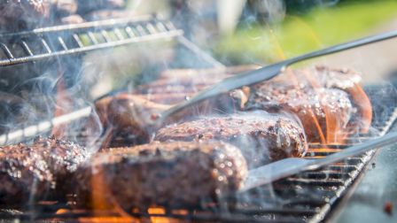 Grilled burgers and sausages on a barbecue with tongs flipping meat, surrounded by smoke and flame, perfect for a summer cookout.