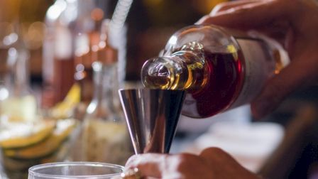 A person pours a brown liquid from a bottle into a jigger, surrounded by bar tools and sliced lemons on a counter.