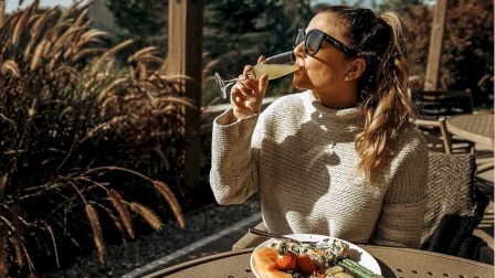 A person in sunglasses is outdoors, enjoying a drink with a plate of food and a cup, surrounded by greenery and sunlight.
