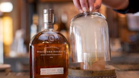 A bottle of Woodford Reserve whiskey and a cocktail being infused with smoke under a glass dome on a bar counter.
