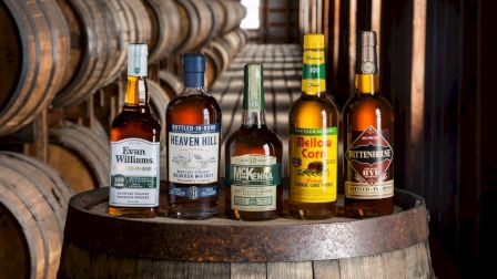 Five whiskey bottles sit on a barrel in a storage area lined with barrels in the background.