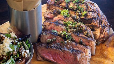 A juicy, sliced steak garnished with herbs on a wooden board, accompanied by a metal container and a salad on the side, sits ready.