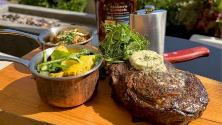 A grilled steak with herb butter, greens, copper pots with side dishes, and a bottle of bourbon on a wooden board.