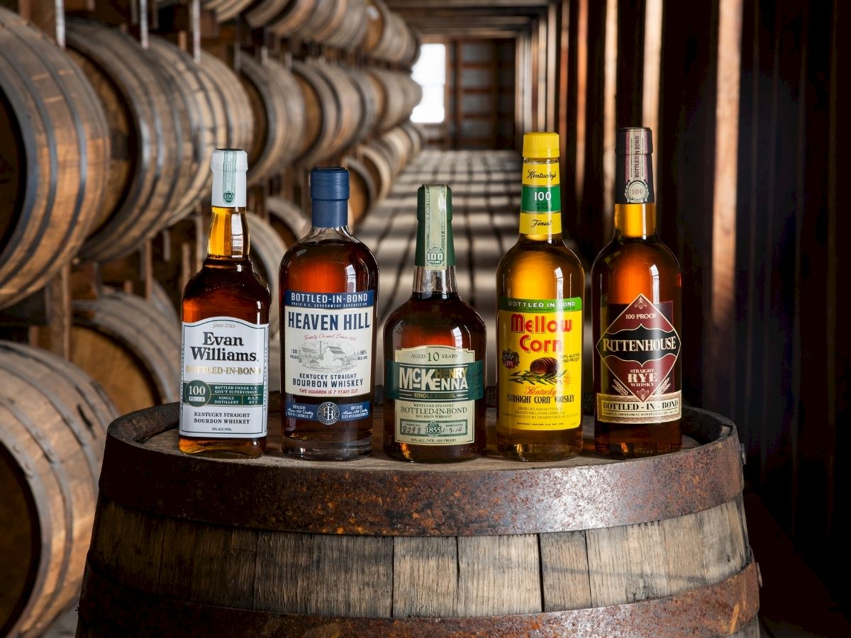 Five bottles of whiskey are displayed on a barrel in a cellar filled with more barrels in the background.