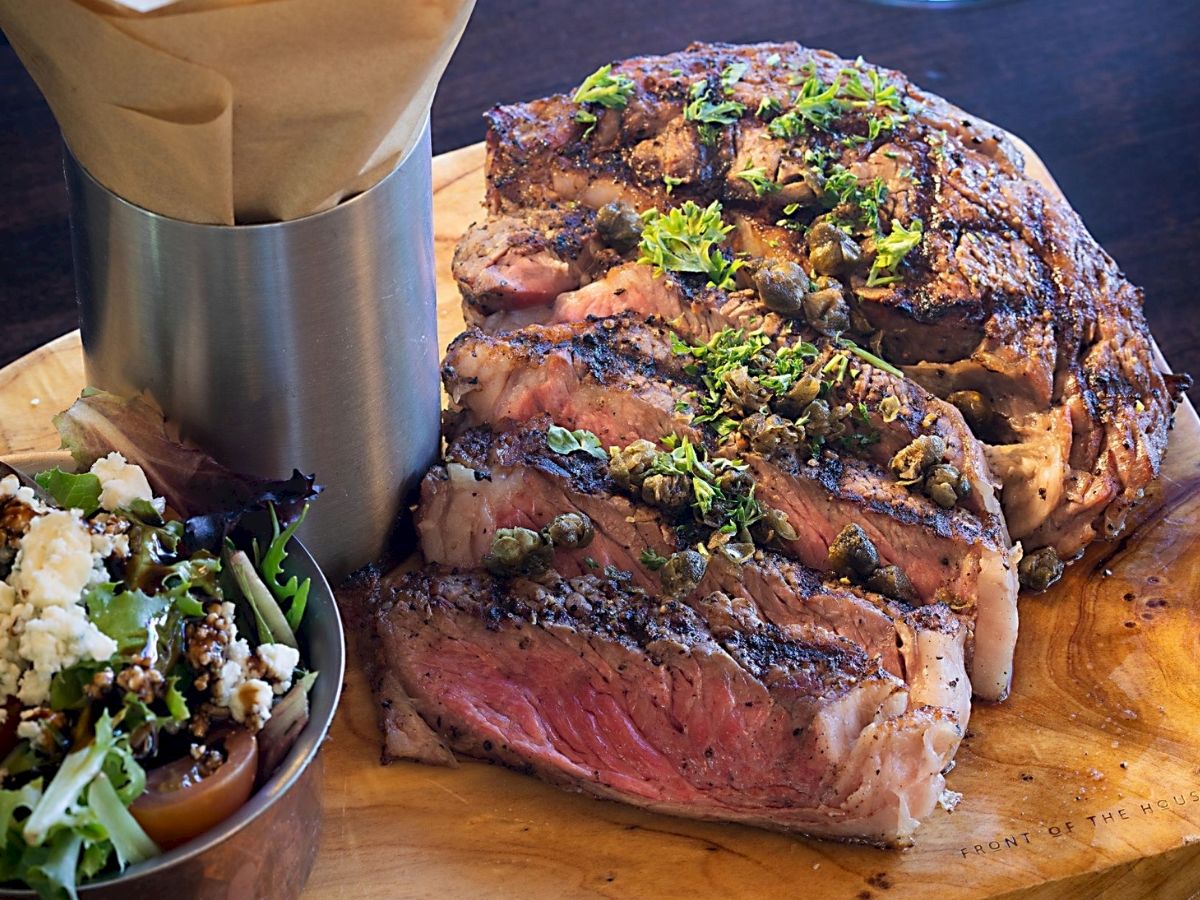 A succulent sliced steak garnished with herbs, accompanied by salad and fries in a metal cup on a wooden board.
