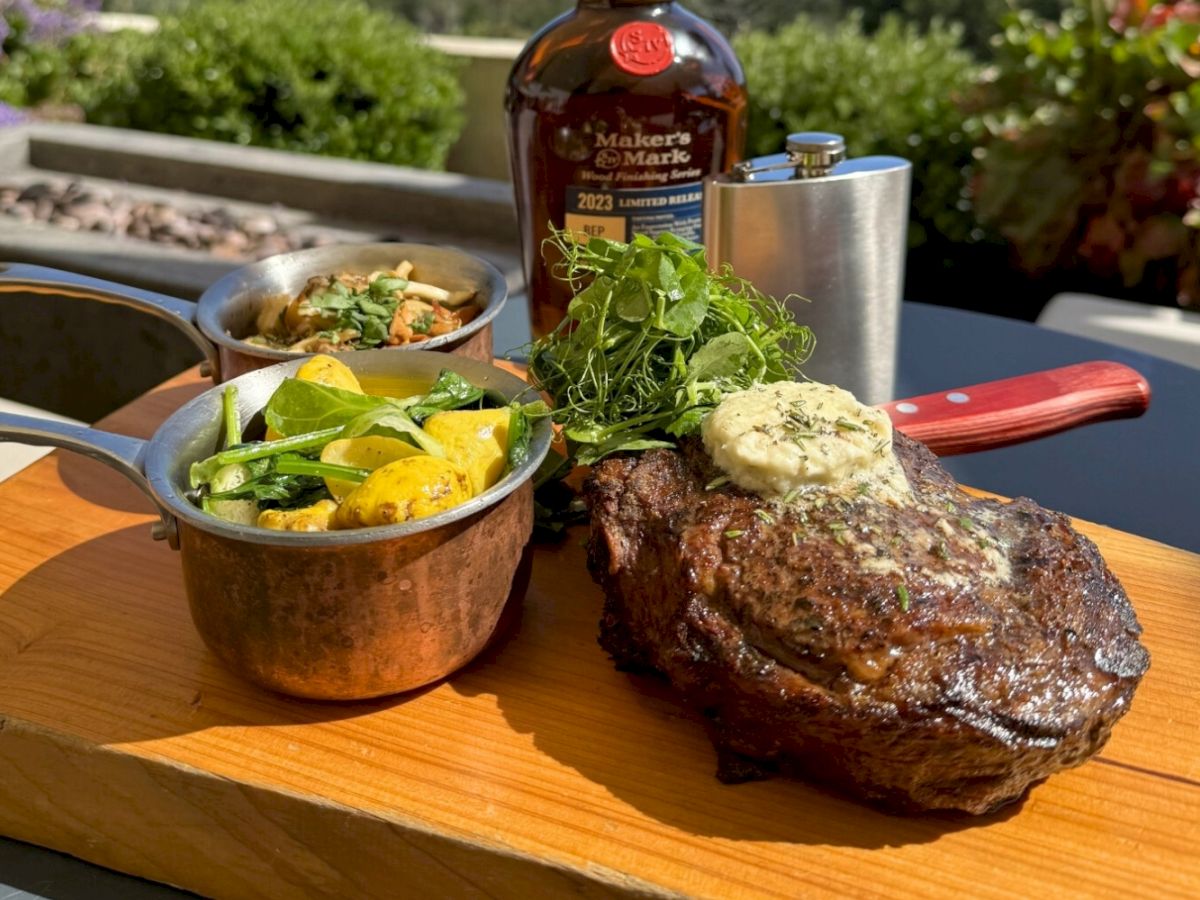 Steak with herb butter, greens, cooked vegetables, and a bottle of Maker's Mark on a wooden board, served outdoors.