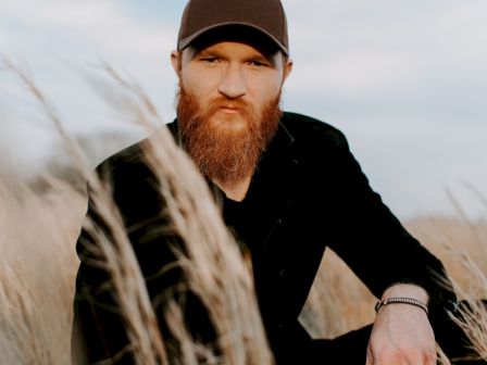 A man with a beard and wearing a dark cap is sitting outdoors among tall grasses against a cloudy sky.