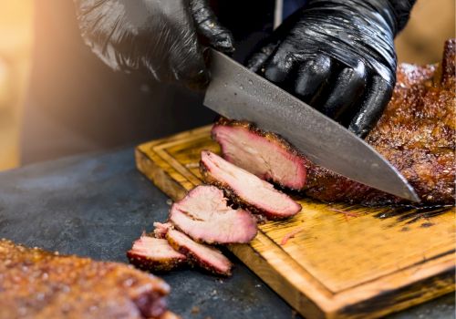 A person wearing black gloves is slicing smoked meat on a wooden cutting board with a knife.